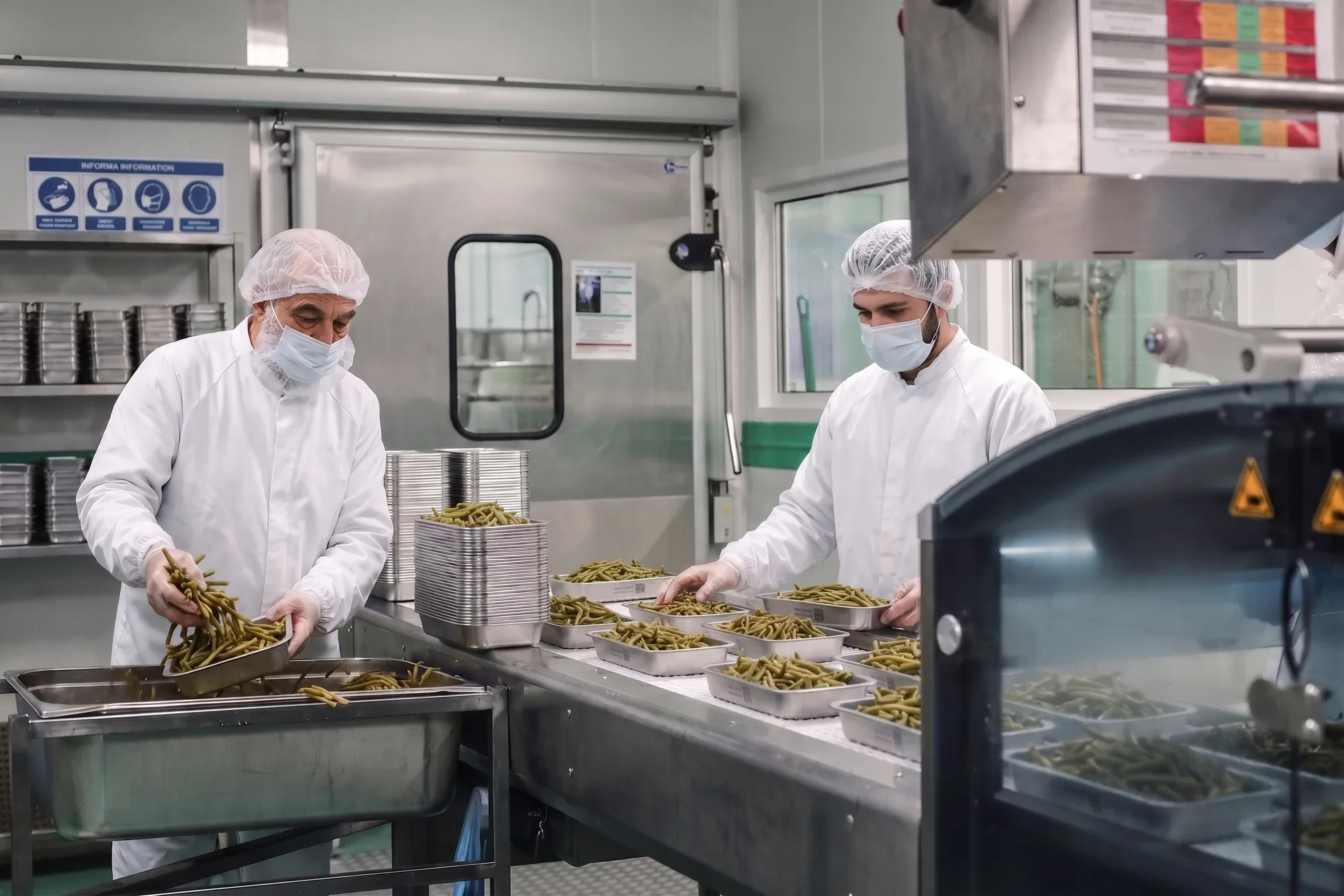 Deux agents portionnent des haricots verts dans des bacs GN inox, sur la ligne d’une cuisine centrale