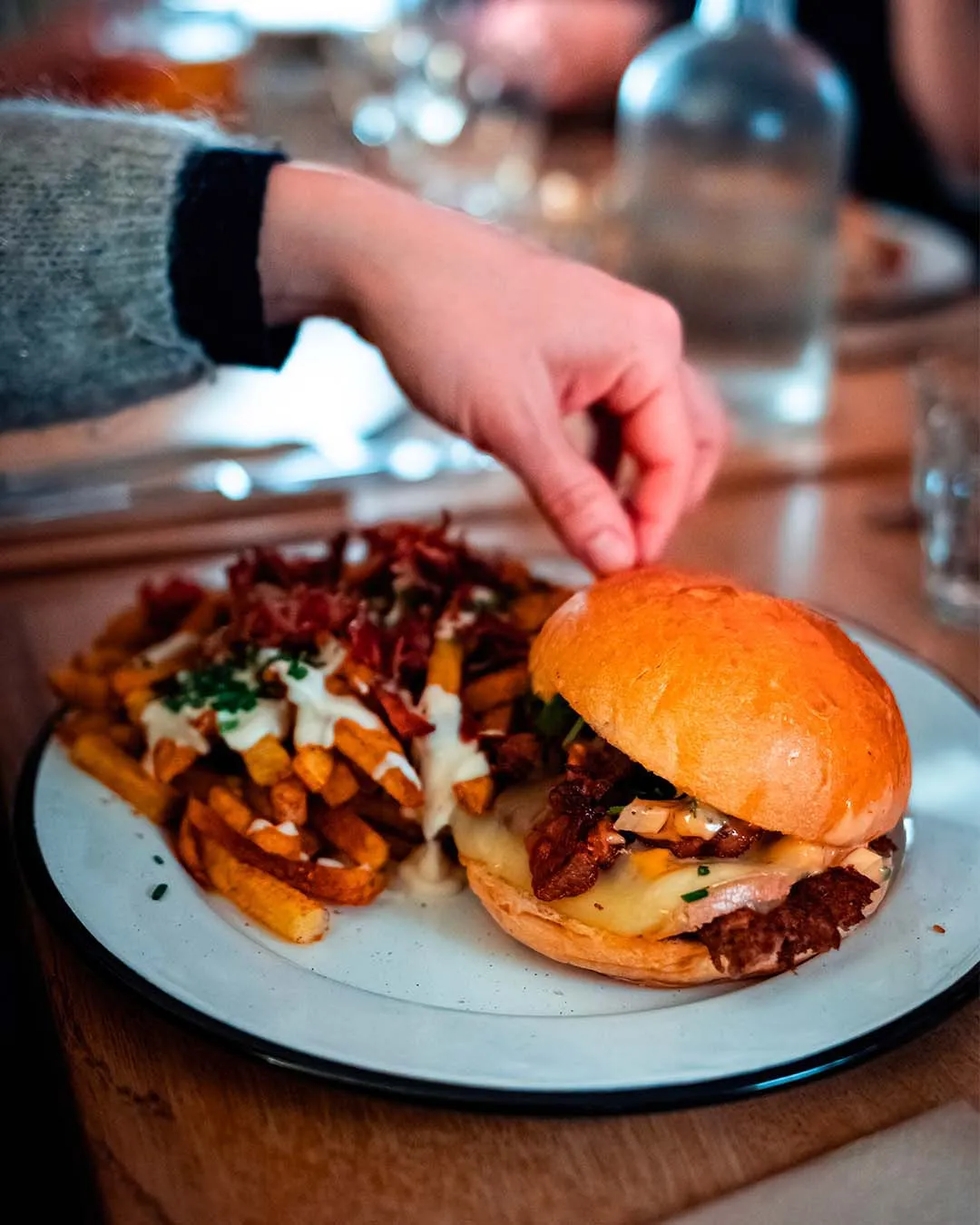 Burger et frites servis dans une assiette émaillée blanche à bordure noire, une main venant saisir le pain