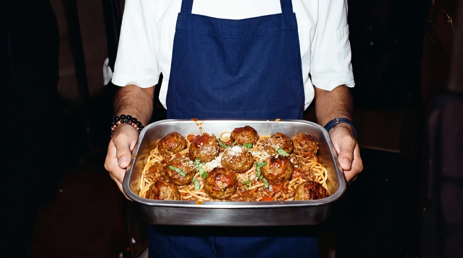 Un cuisinier en tablier bleu présente à deux mains un plat inox de spaghettis aux boulettes de viande, parmesan et basilic