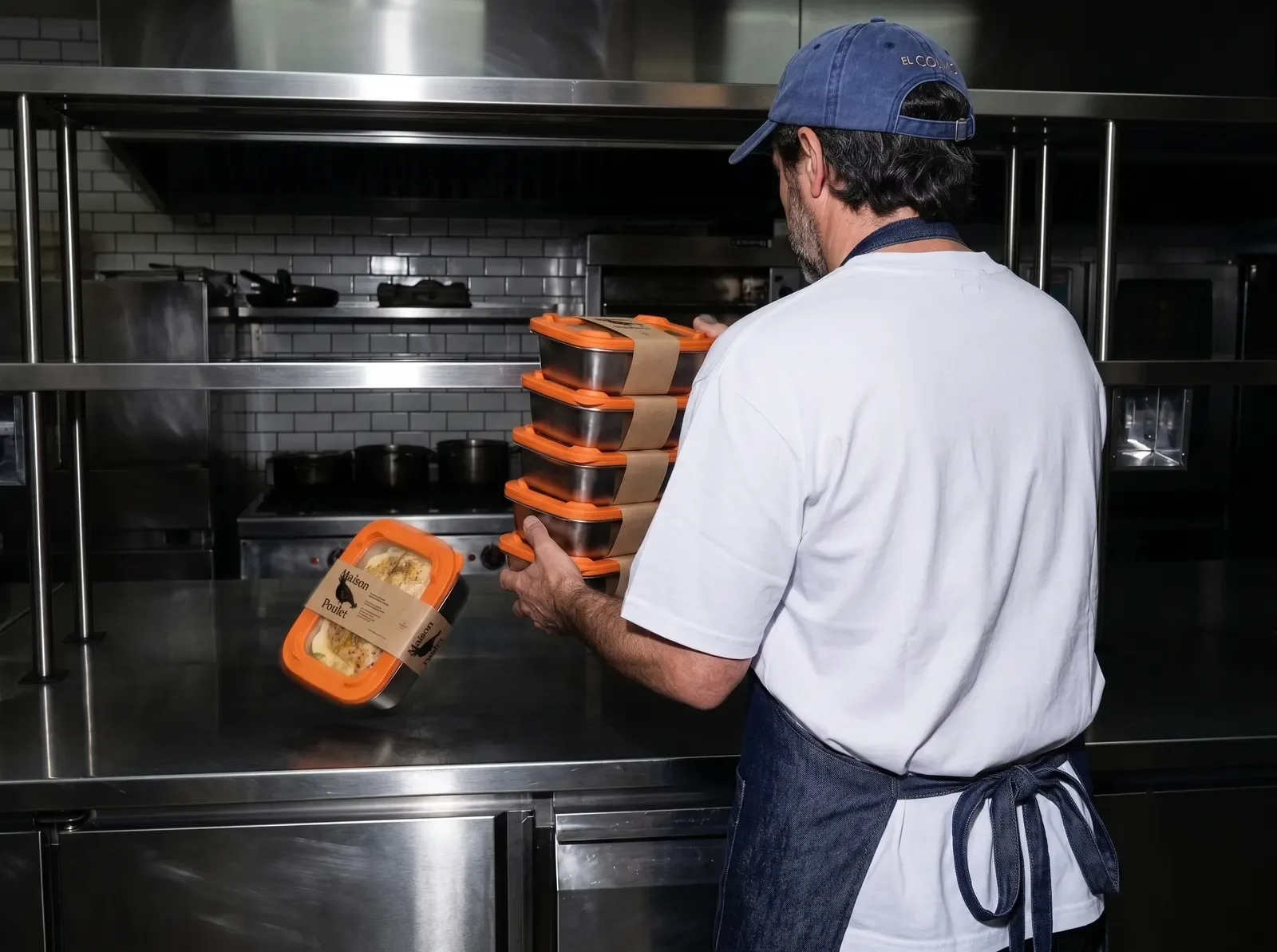 Un cuisinier emporte une pile de quatre barquettes inox à couvercle orange, ceinturées de bandes kraft, dans une cuisine professionnelle