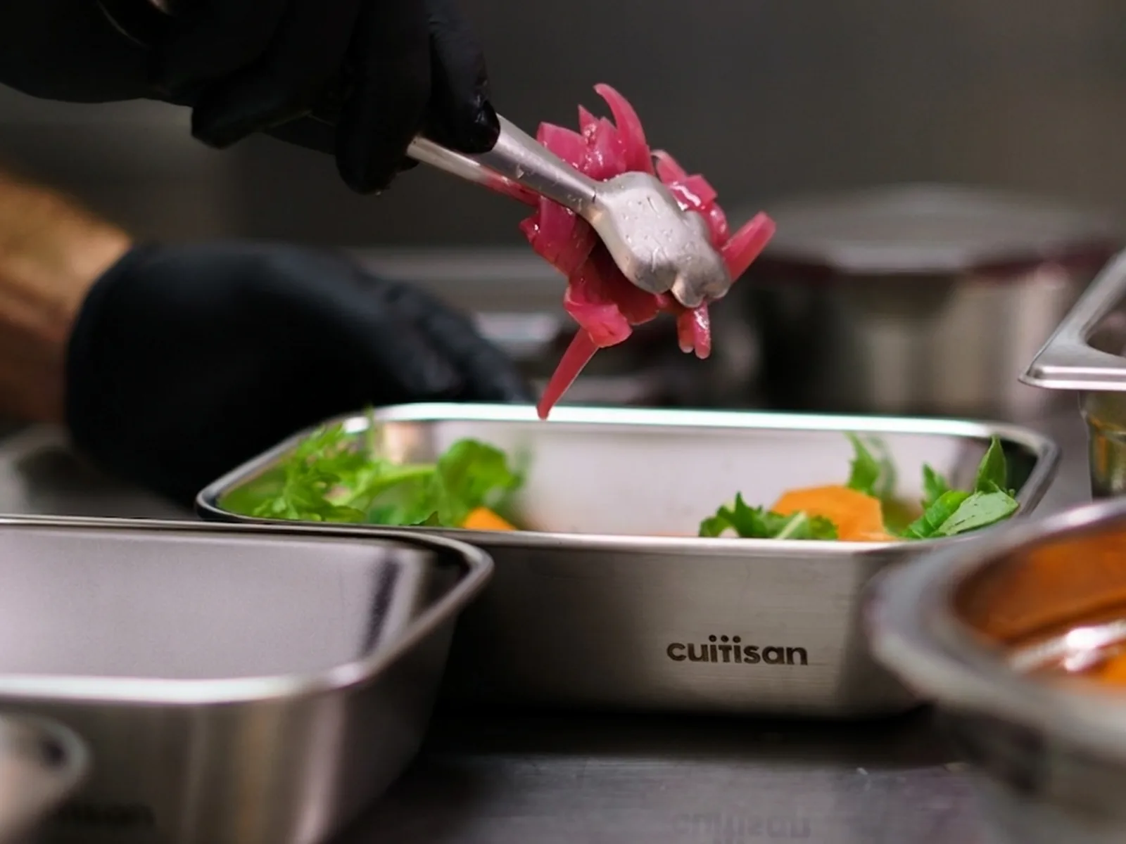 Un cuisinier dresse des oignons rouges marinés à la pince dans une boîte inox Cuitisan